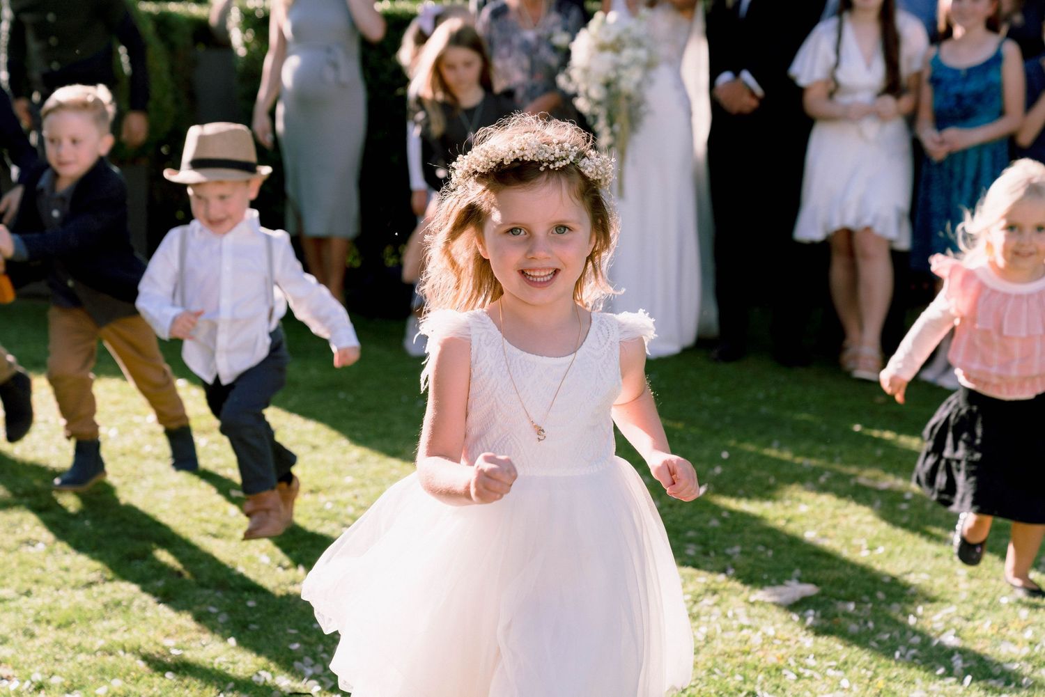 A child in a white flower girl dress runs joyfully across a sunny lawn during an outdoor wedding celebration.