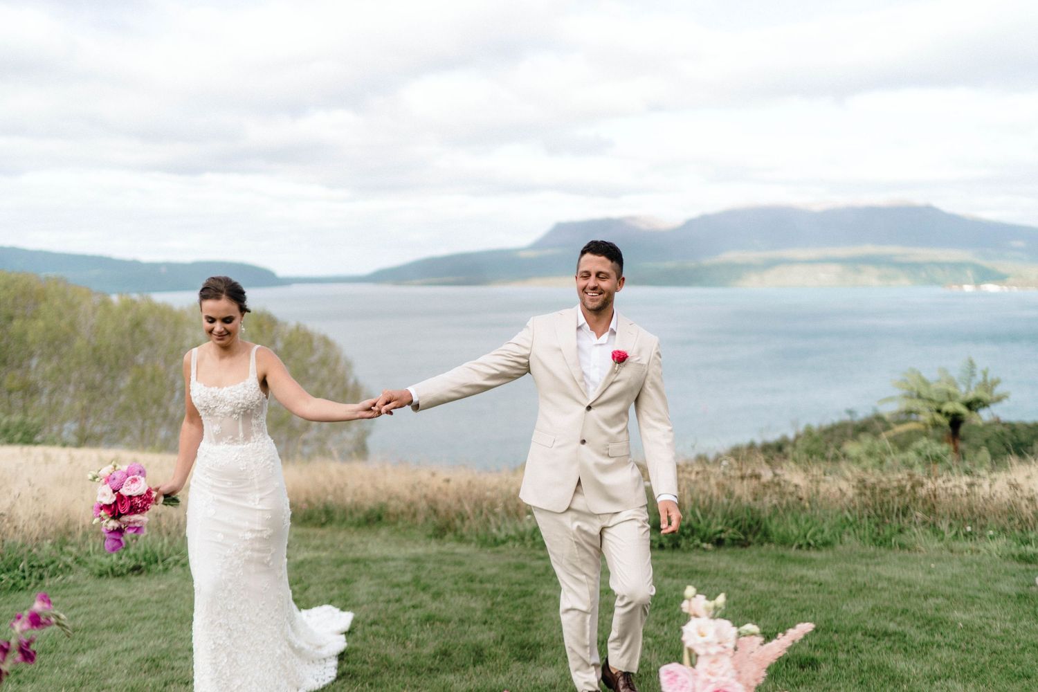 Wedding couple holds hands while standing on grassy cliffside overlooking a scenic lake and mountains.
