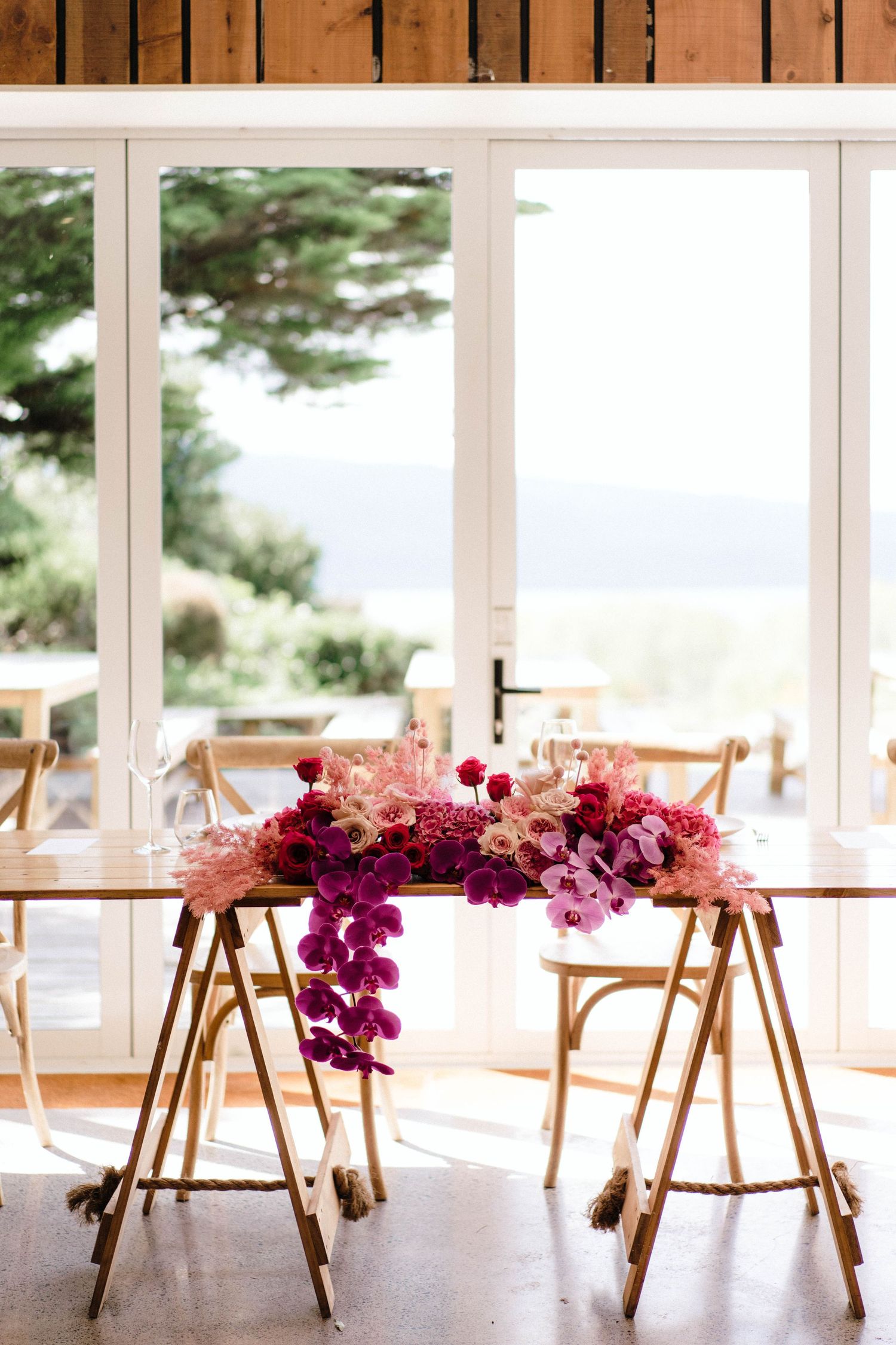A stunning floral arrangement with pink and purple blooms cascades over a table with gold metal sawhorse legs near glass doors.