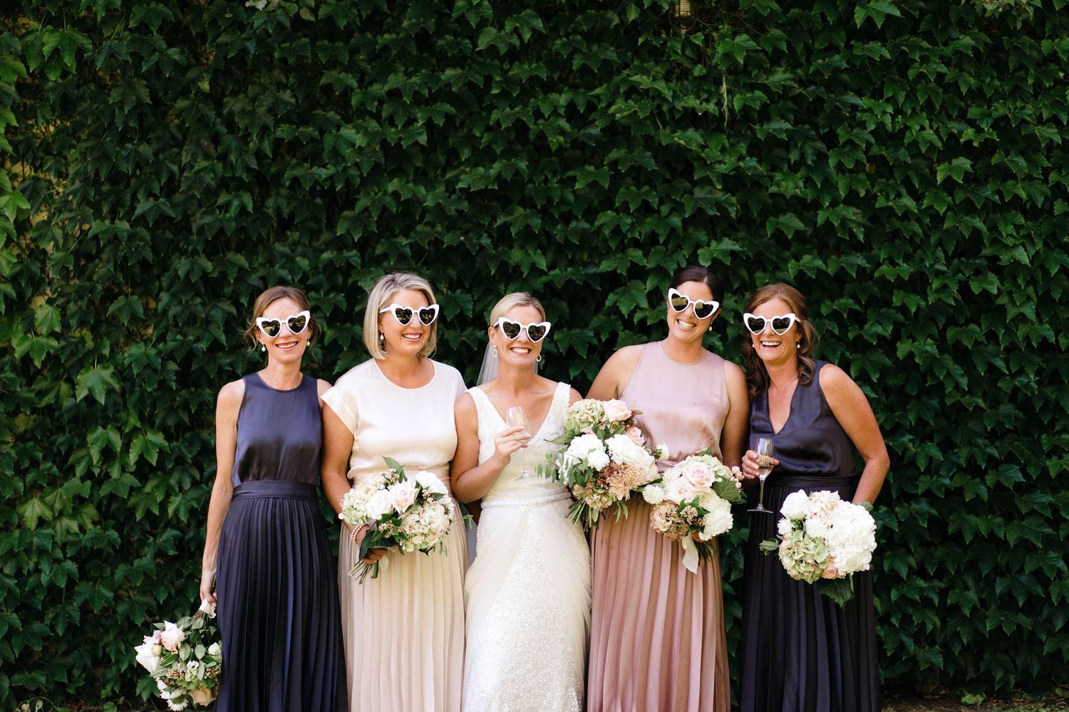 Wedding party in navy and blush dresses wearing sunglasses pose with bouquets against an ivy wall.