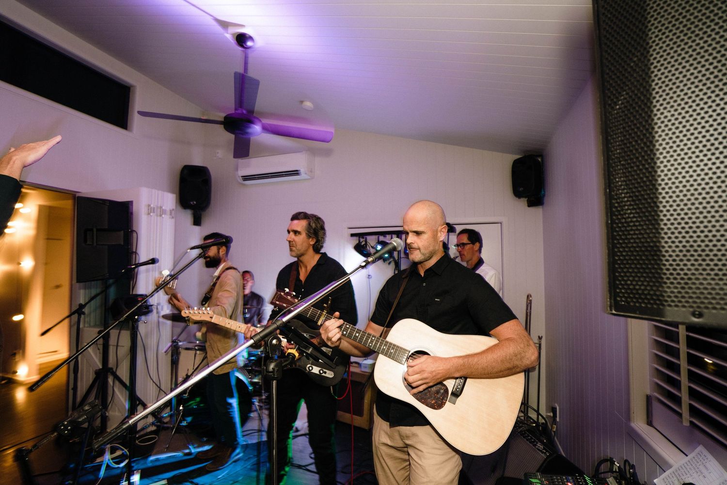 A live band performs on stage with guitars and microphones under purple lighting at an indoor venue.