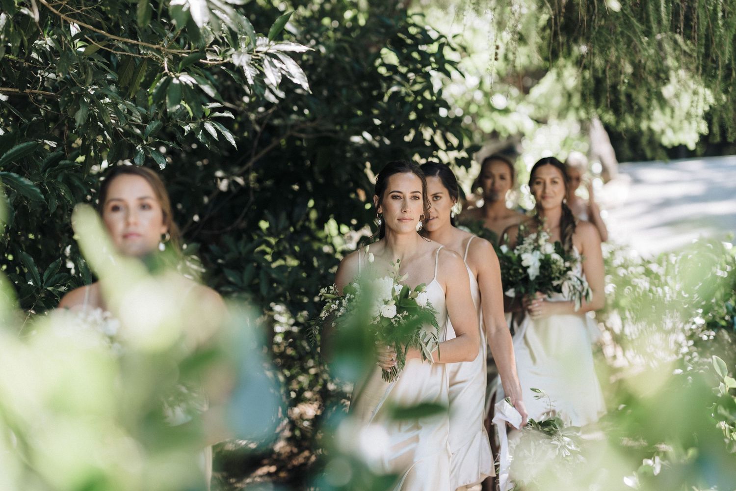 Bridesmaids walking through lush garden pathway carrying bouquets.
