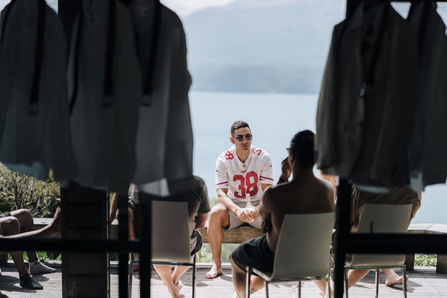 Person in a sports jersey sits in a chair with dark-clad figures in the foreground and a scenic lake view behind them.