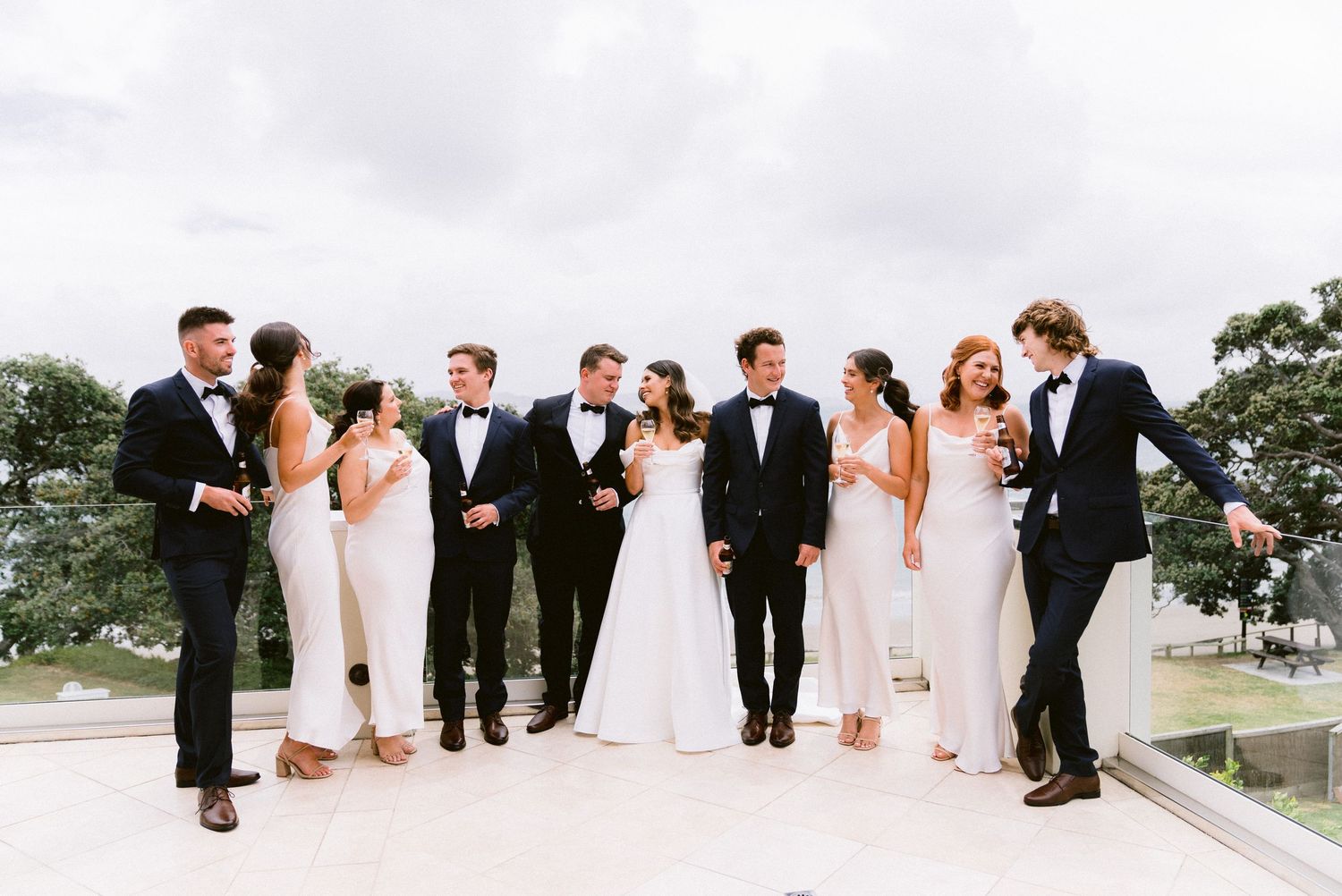 A wedding party in black and white attire poses together on a rooftop terrace against a cloudy sky.