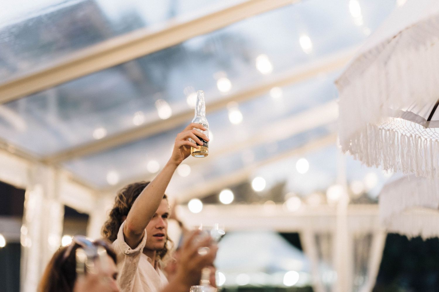 Wedding guests raise glasses in celebration under illuminated tent ceiling.