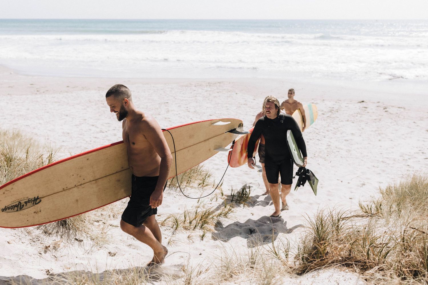 Surfers walking along a sandy beach carrying surfboards on a sunny day.