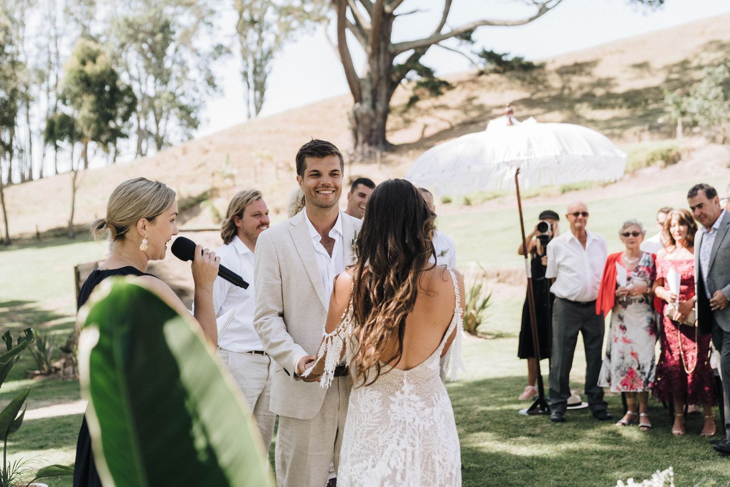Outdoor wedding ceremony taking place under trees with guests gathered on a sunny day.