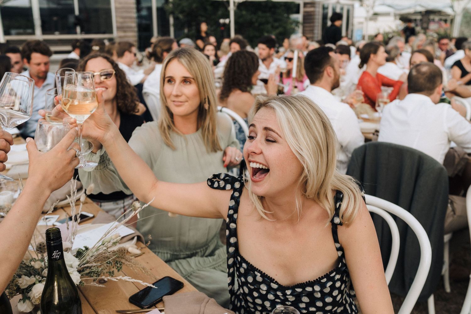 Wedding guests laughing and raising glasses during an outdoor reception celebration.