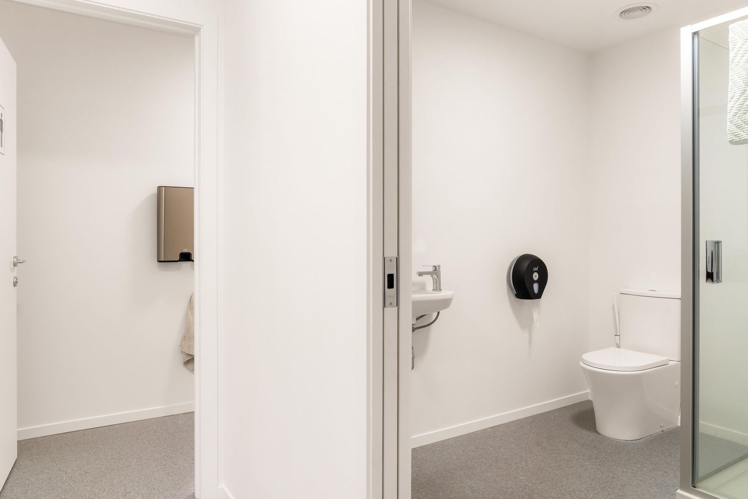 Contemporary office restroom with sleek white fixtures, glass partitions and clean minimalist design.