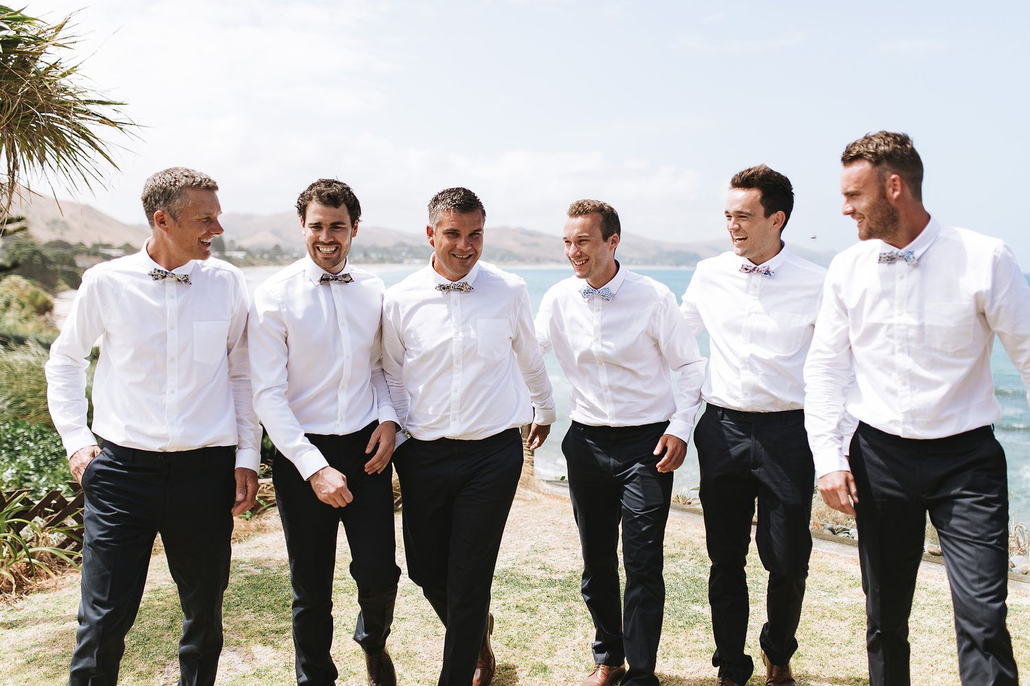 Groomsmen in matching white shirts and dark pants stand together on a beach boardwalk.