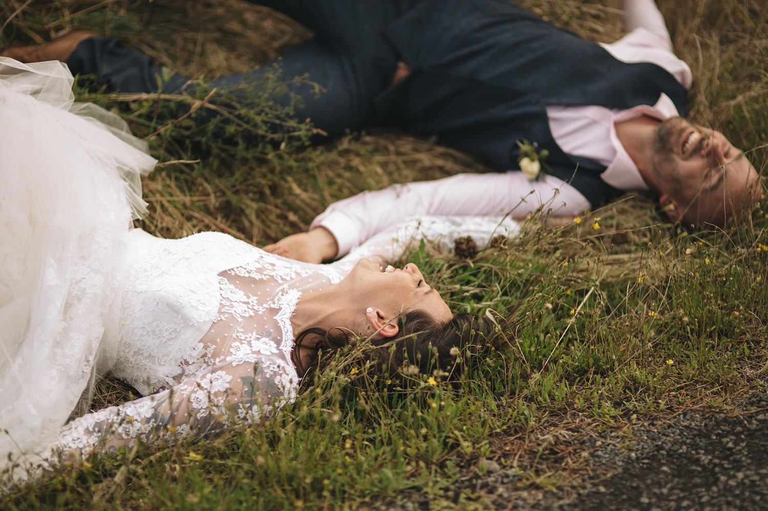 A couple in wedding attire lies together on grass in a romantic outdoor setting.