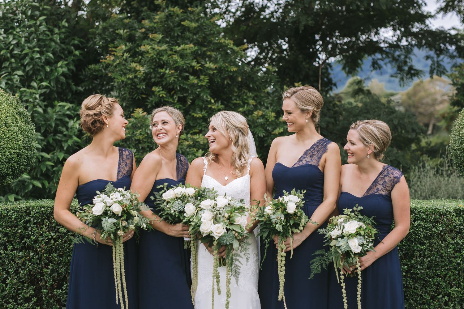 Four bridesmaids in navy blue dresses hold elegant white and green wedding bouquets while facing a lush garden backdrop.