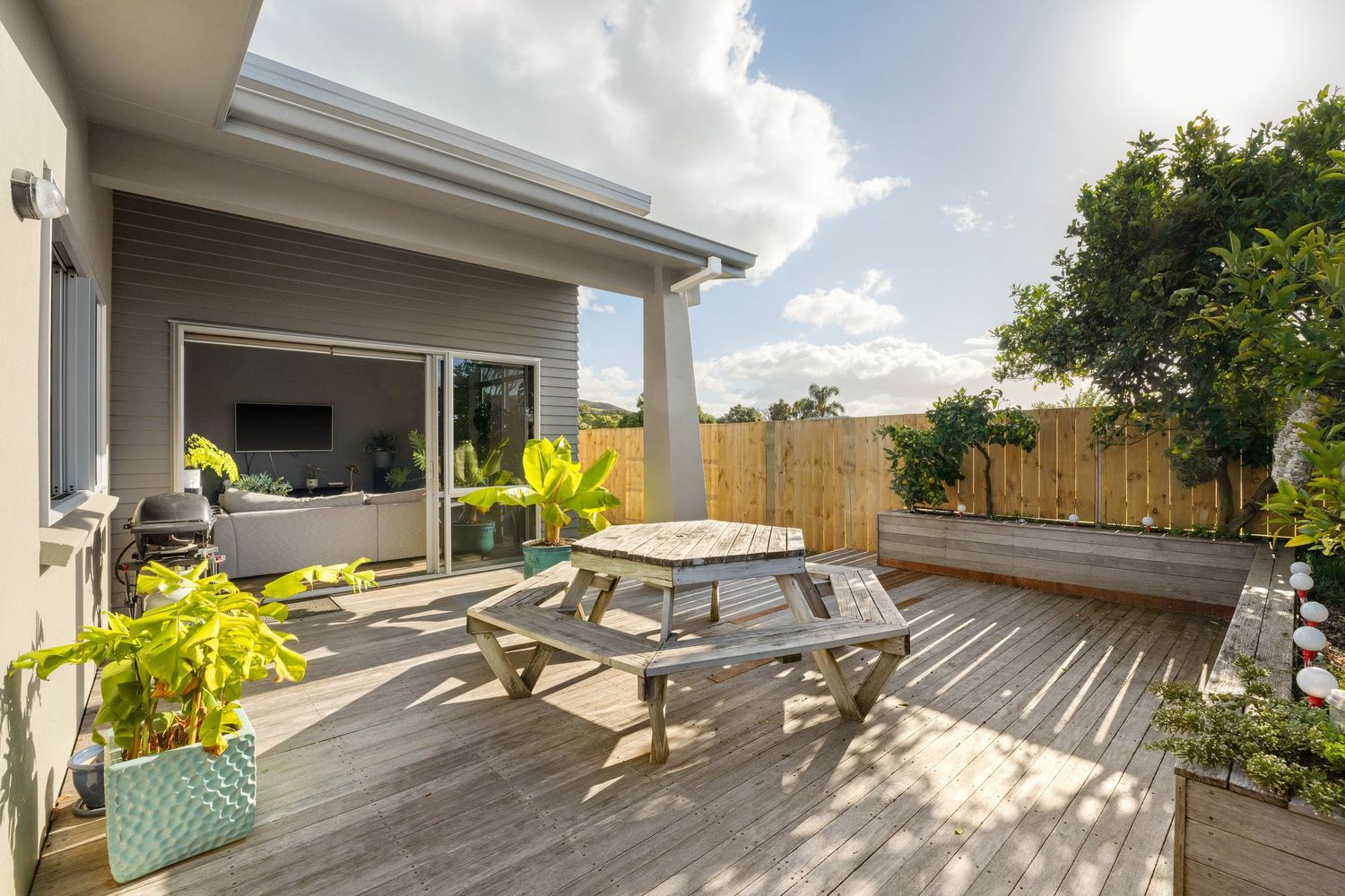 Modern backyard deck with covered patio area featuring a picnic table and lush greenery under bright blue skies.