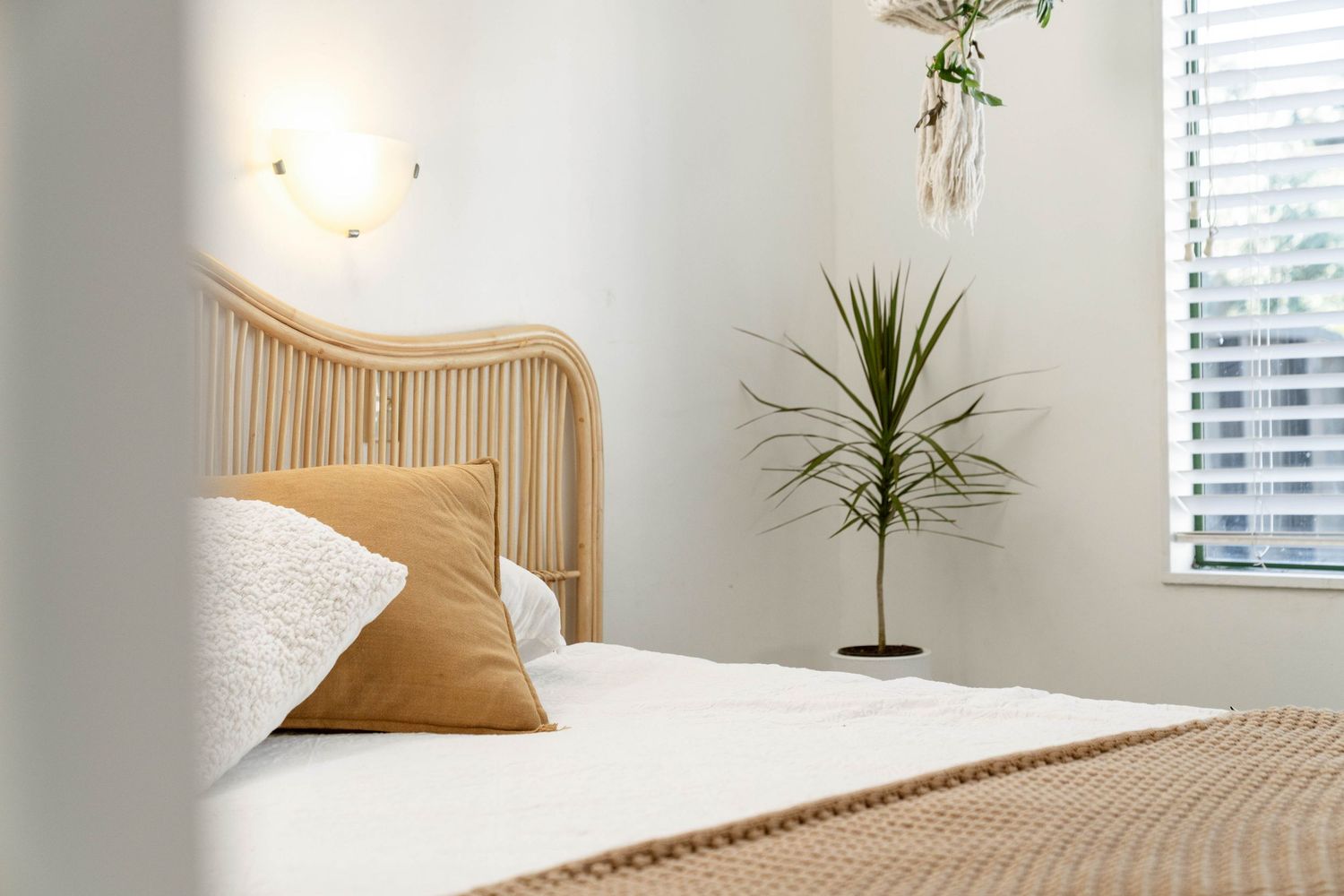 Cozy bedroom with wooden headboard, white bedding, tan throw blanket and potted plant next to window.