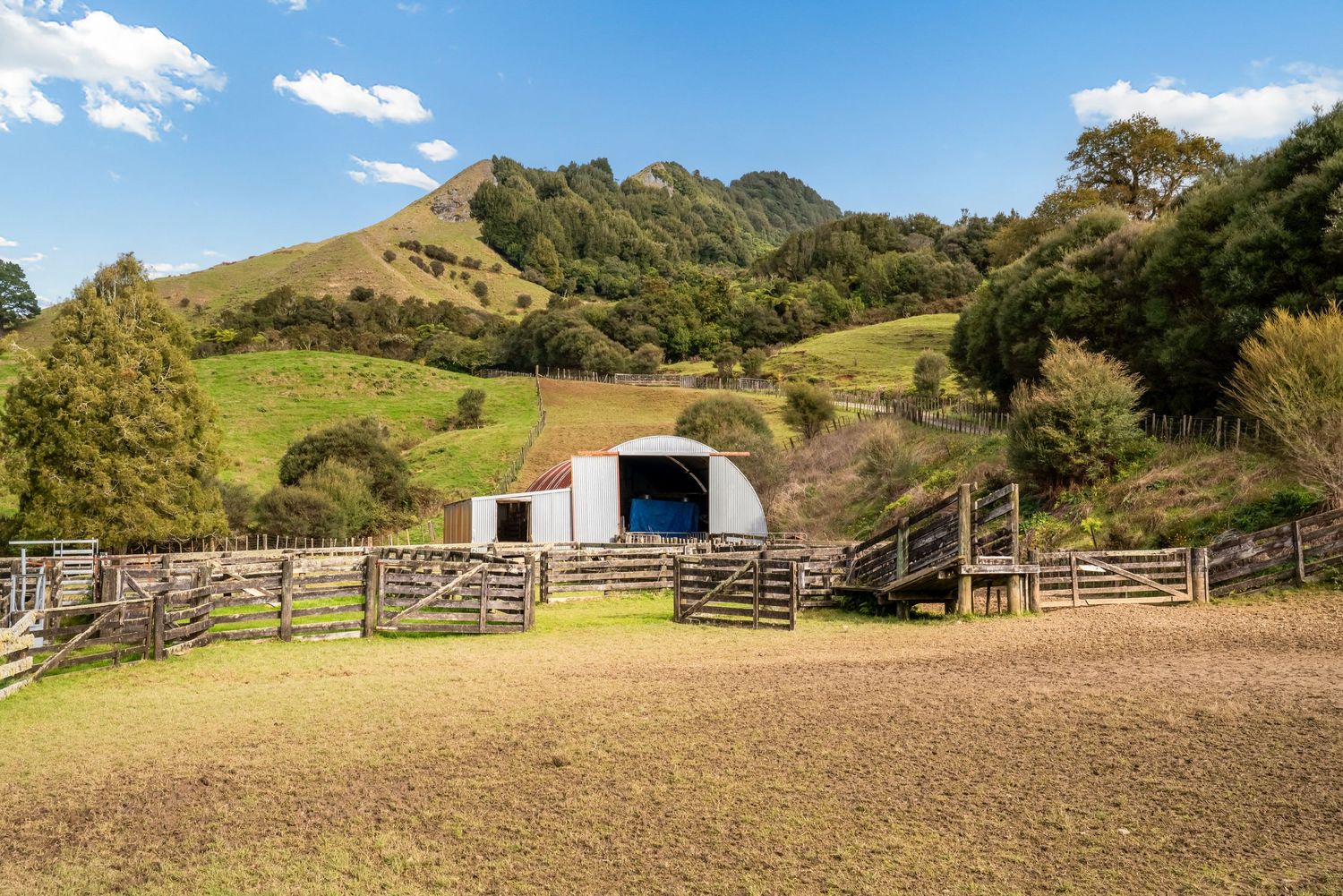 Small barn with wooden fence and paddock nestled in rolling green hills under blue sky.