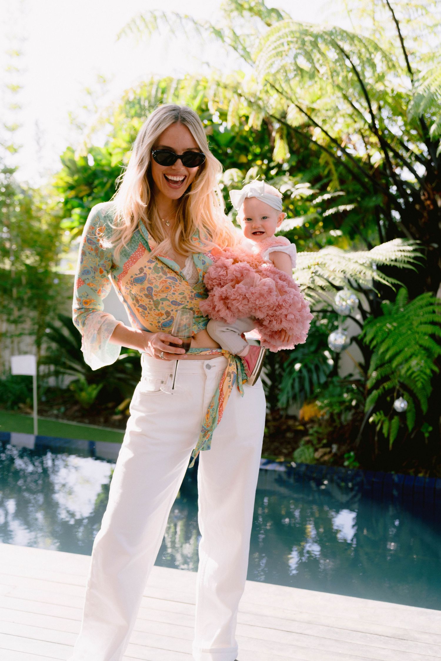 Stylish summer portrait by pool with baby in pink dress and woman in white pants.