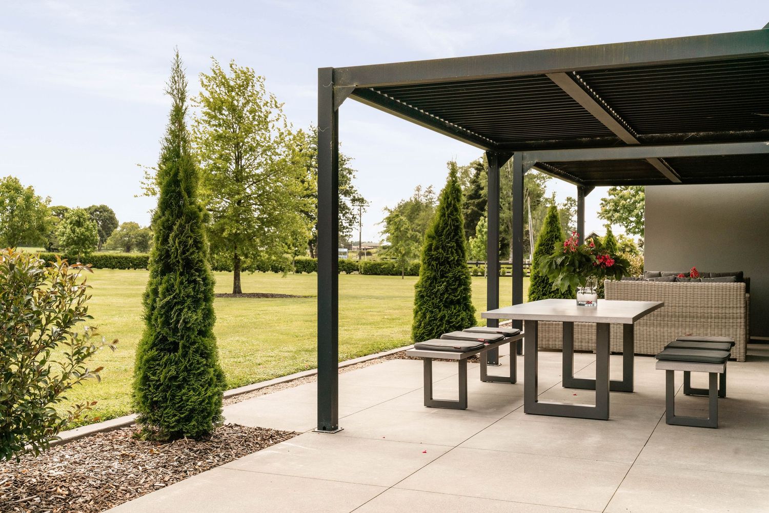 Modern outdoor patio area with contemporary furniture and tall cypress trees overlooking a spacious green lawn.