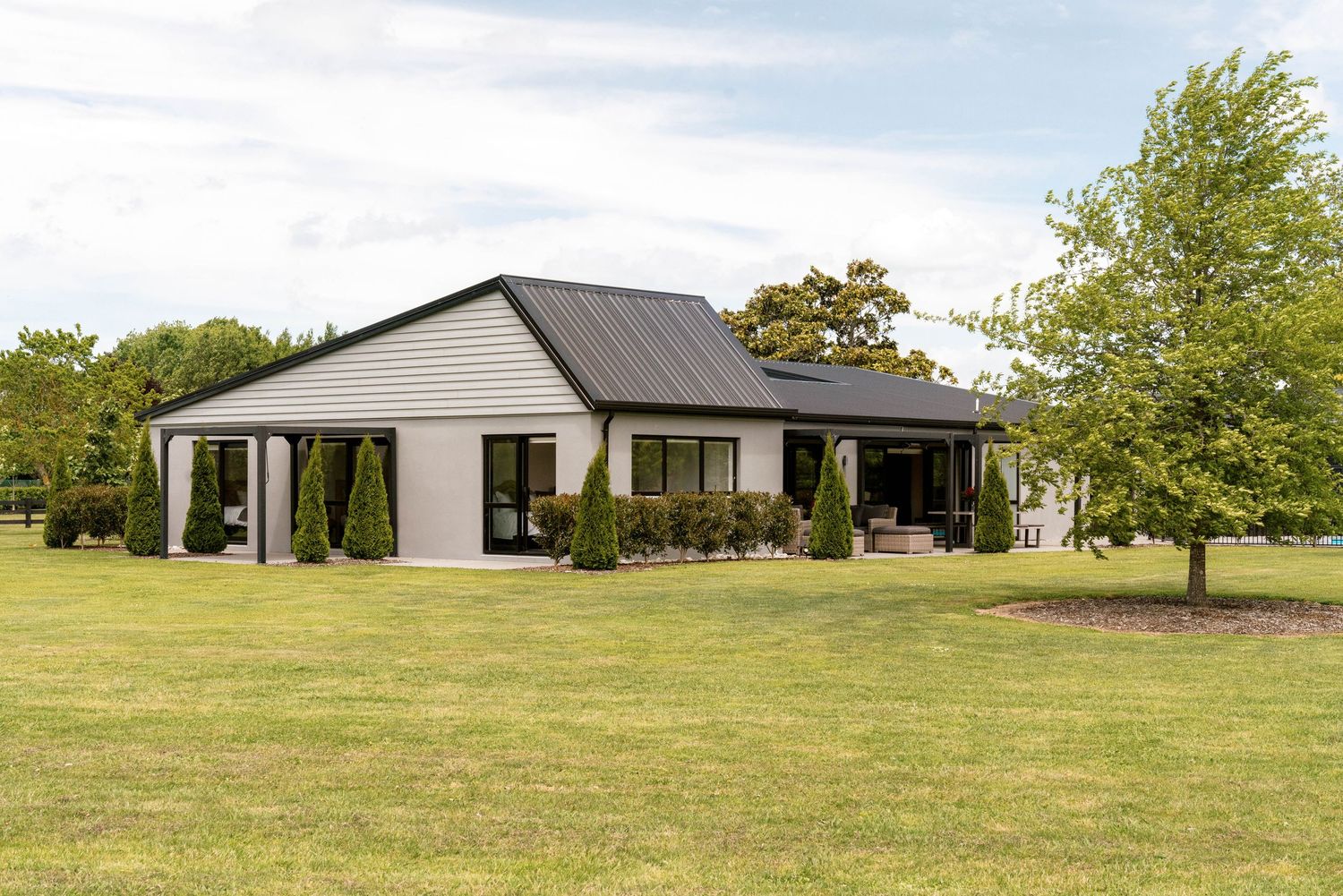 Modern single-story home with dark roof and white exterior sits on a spacious green lawn surrounded by trees.