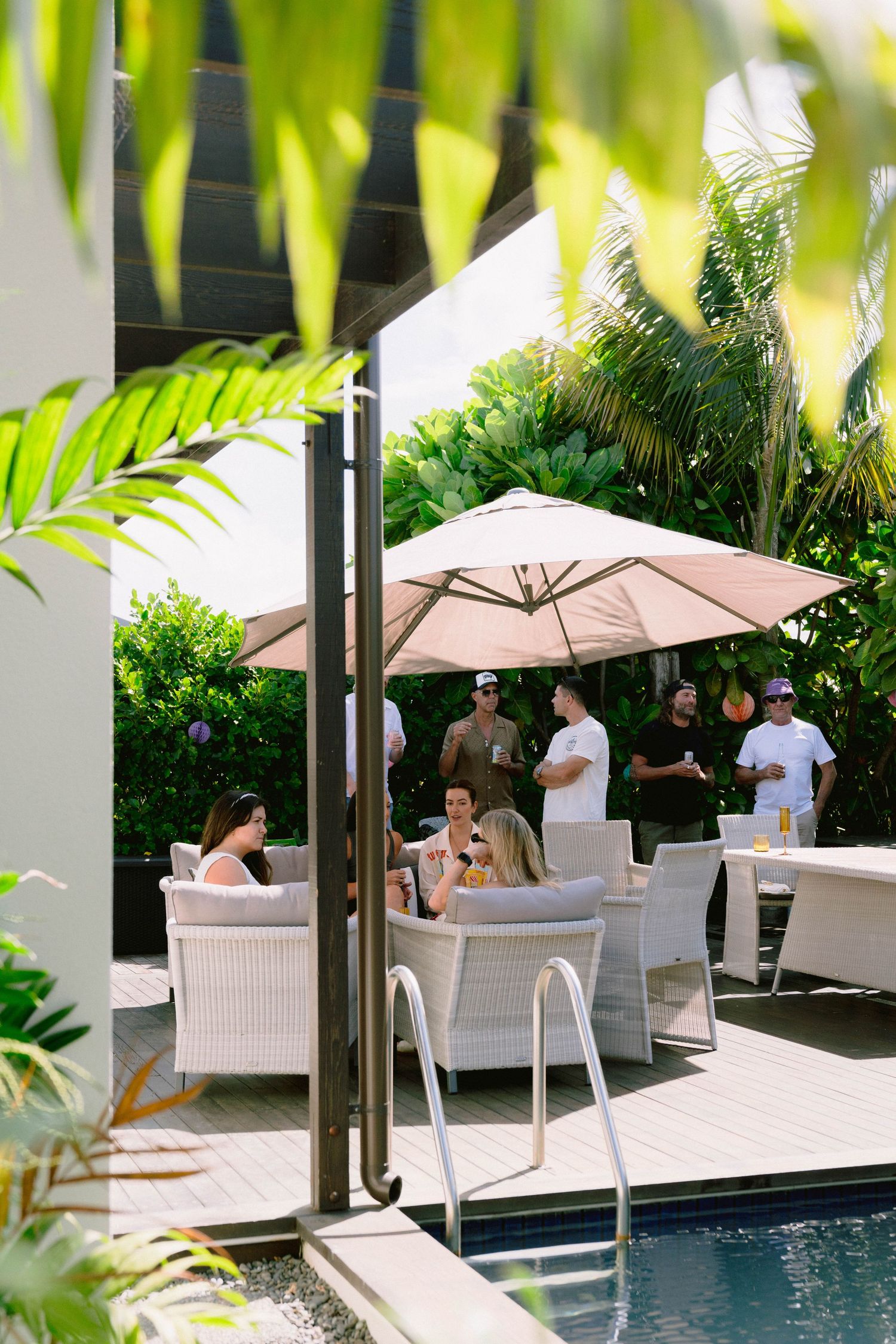 People relax by a pool under white umbrellas with tropical plants and palm fronds creating a luxurious resort atmosphere.