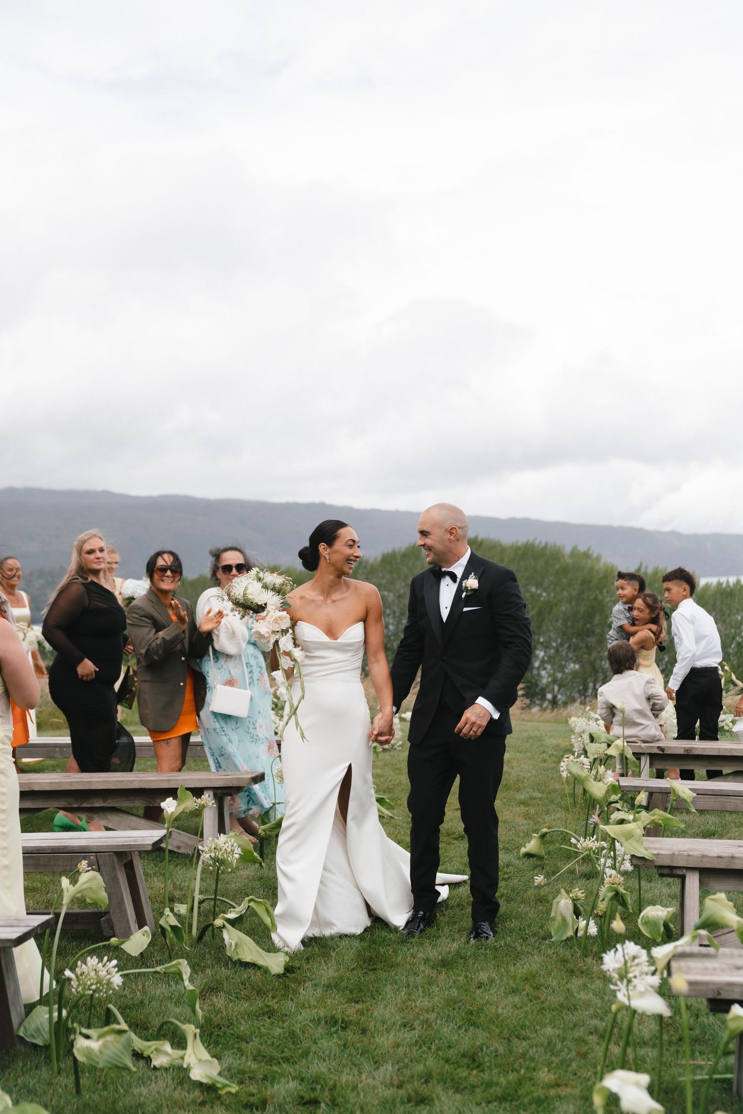 A wedding party gathers on a scenic hillside with rustic picnic tables and white floral decorations.