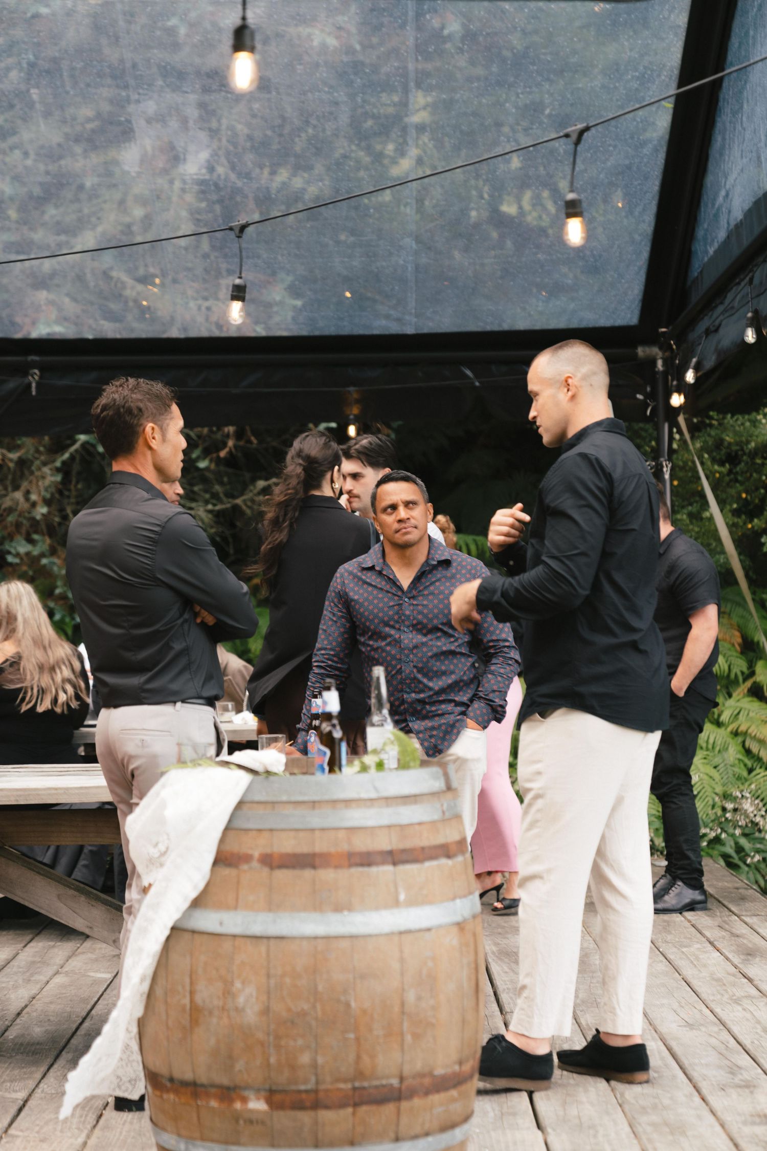 Wedding guests sit and socialize around rustic wooden barrels at an outdoor reception venue under string lights.