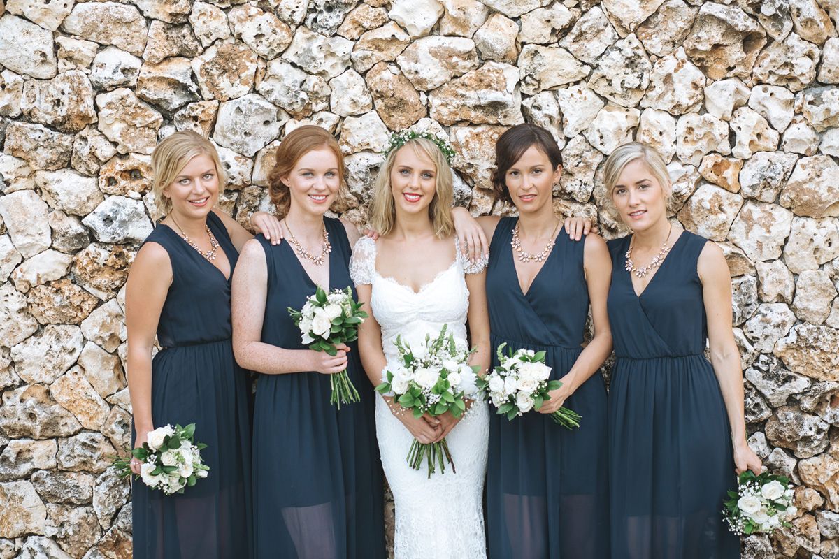Wedding party poses against stone wall with bride in white and bridesmaids in navy blue dresses.