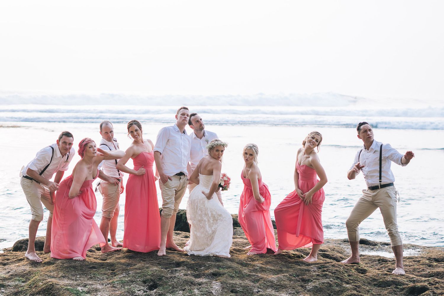Wedding party poses playfully on a beach with ocean waves in the background.