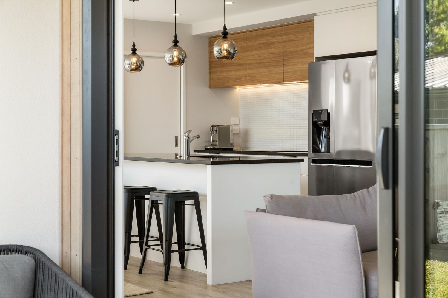 Modern kitchen interior with stainless steel appliances, wooden cabinets, pendant lights and black bar stools at white counter.