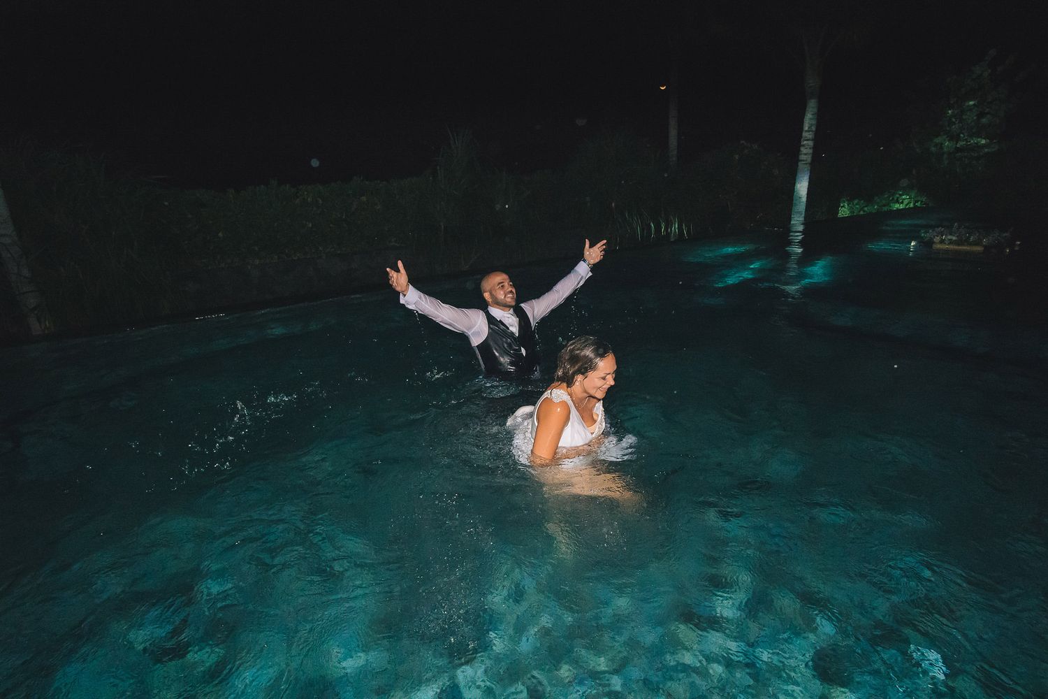 Wedding couple taking a playful nighttime swim in an illuminated pool.