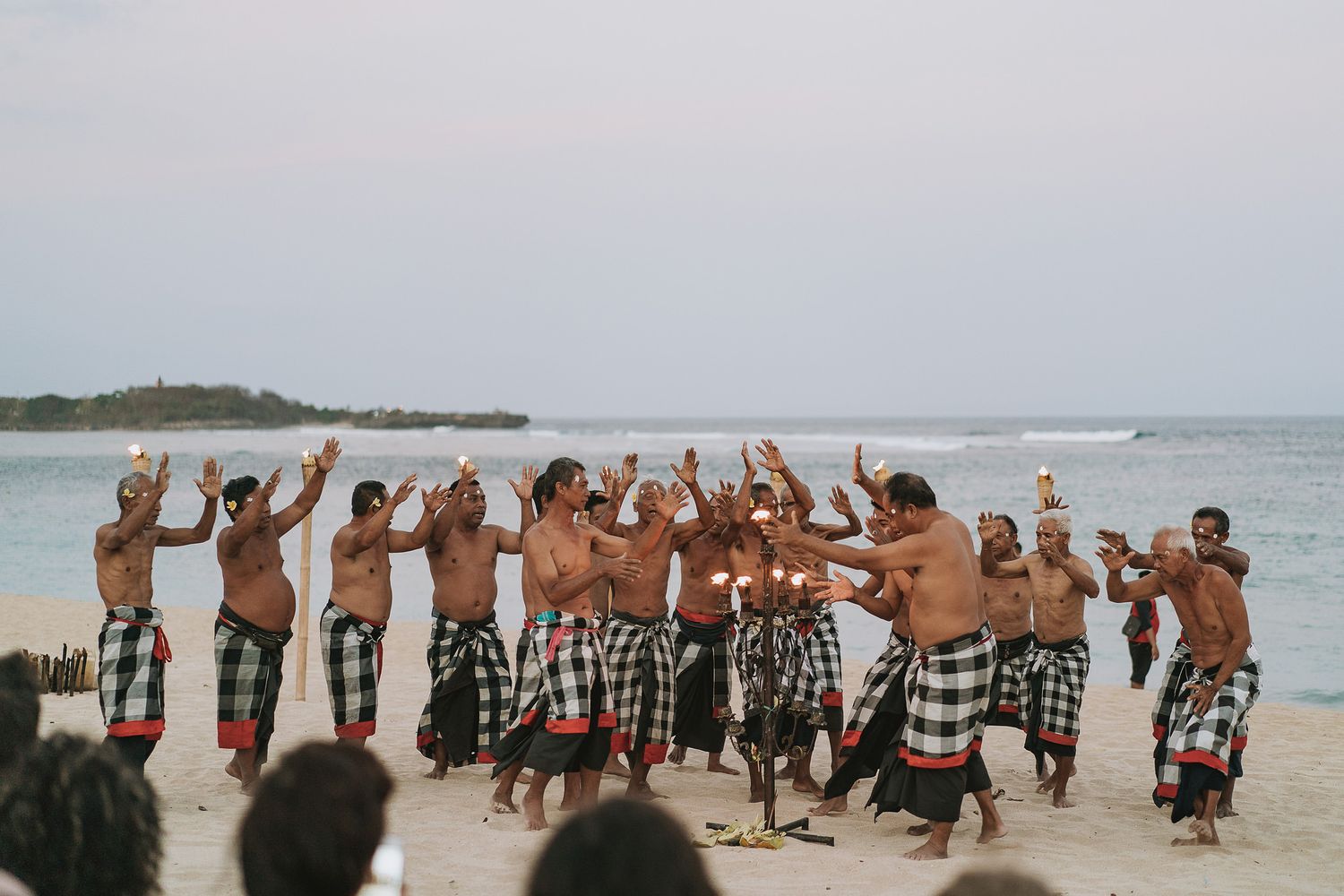 Large group of people performing a traditional Balinese dance on a scenic beach at sunset.
