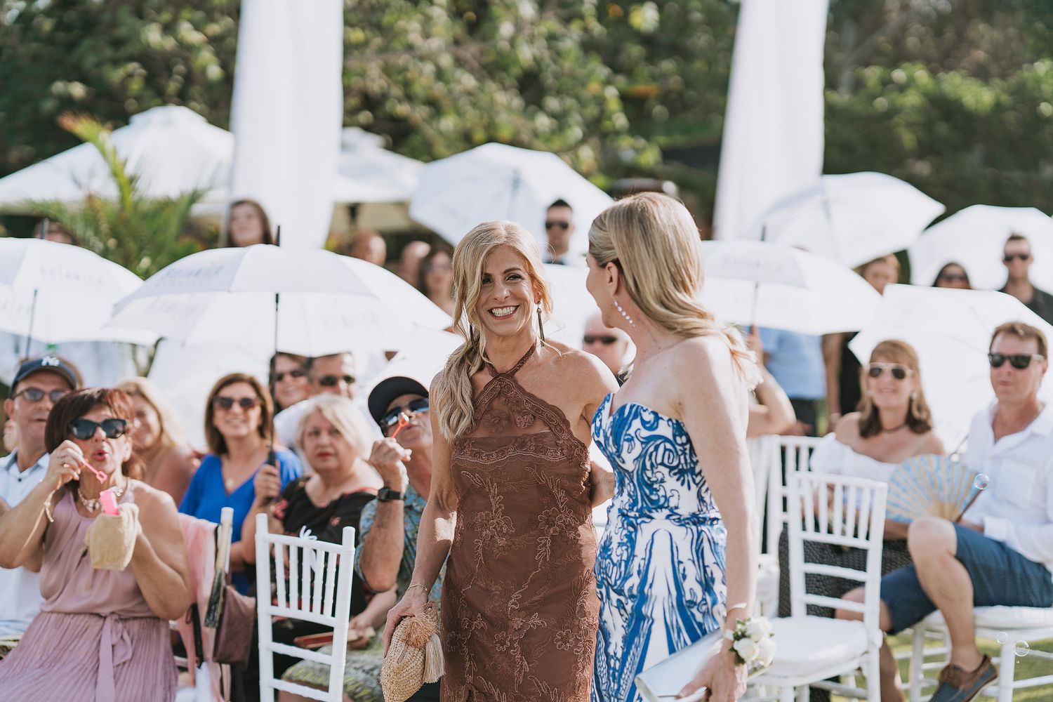 Wedding guests seated outdoors under white umbrellas during daytime ceremony.