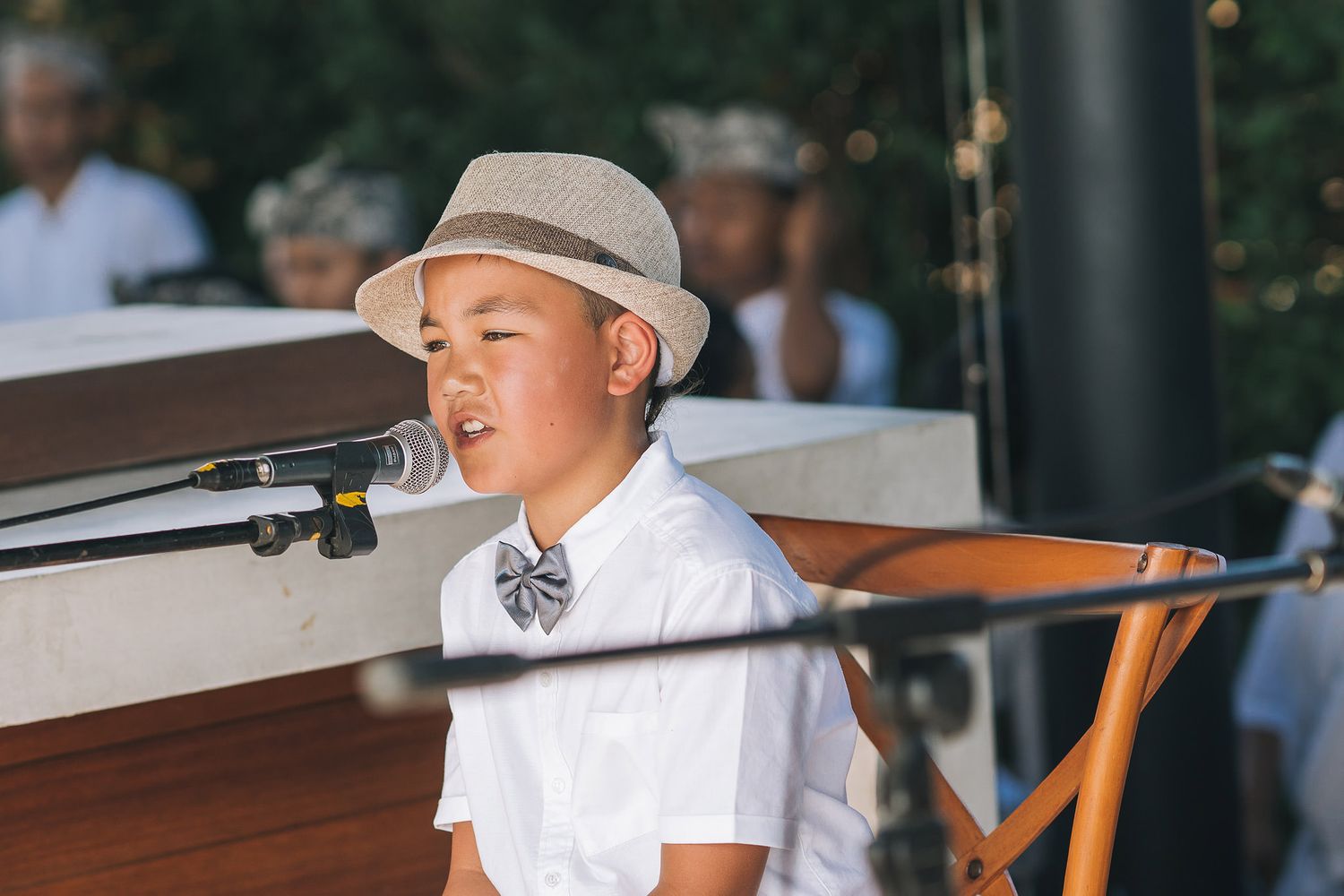 Young performer in white dress shirt and hat singing into microphone on stage during event.
