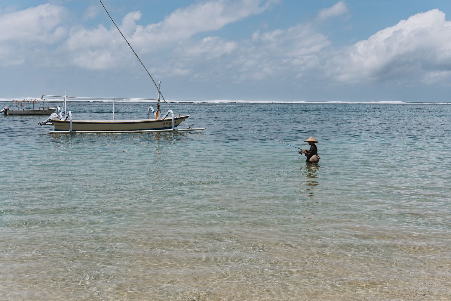 A person wades in shallow tropical waters near a moored fishing boat on a calm sunny day.