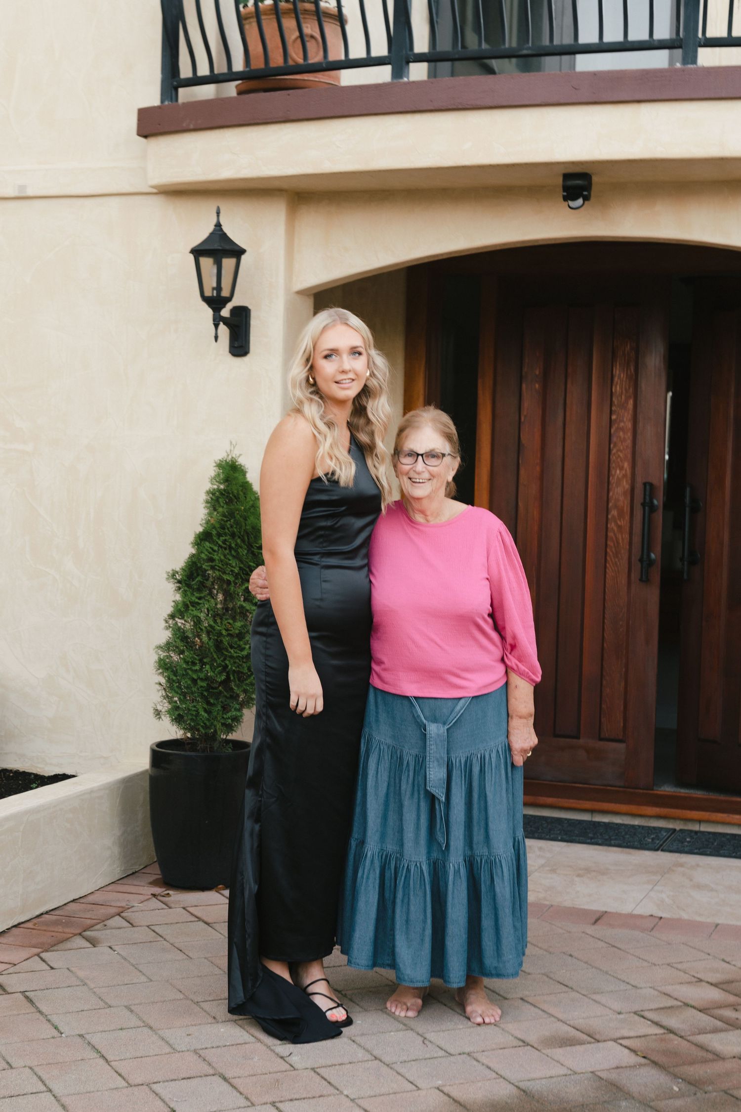 A woman in a black evening dress posing with an older woman in pink at a home entrance.