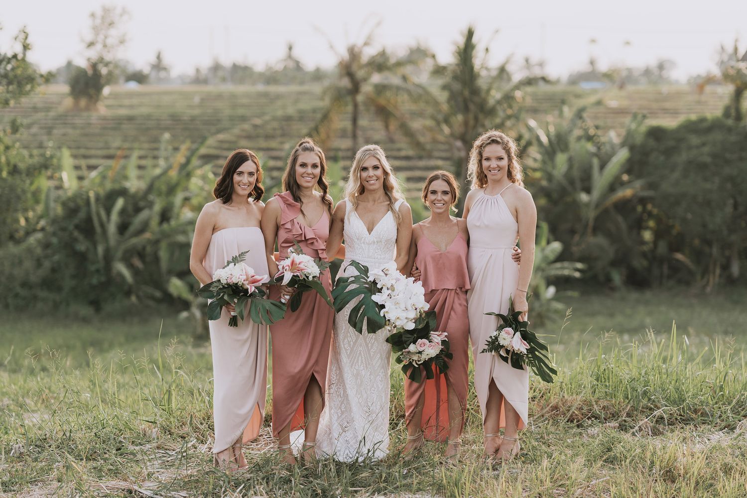 Bridesmaids in pink and blush tones holding tropical bouquets pose together in a lush outdoor setting.