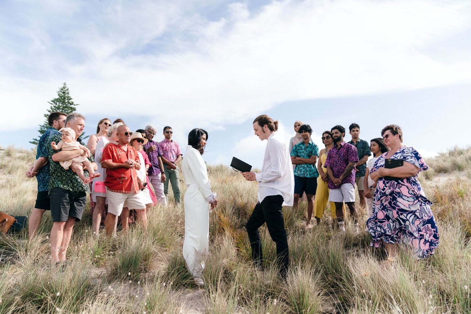 Wedding guests mingling on grassy coastal hillside with ocean view in background.
