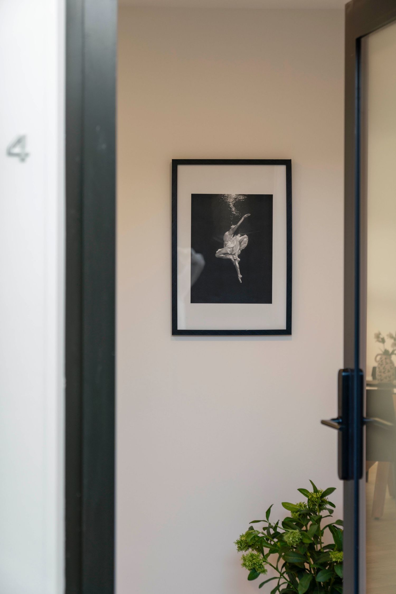 Black and white framed art print hanging on beige wall next to green plant and door handle.