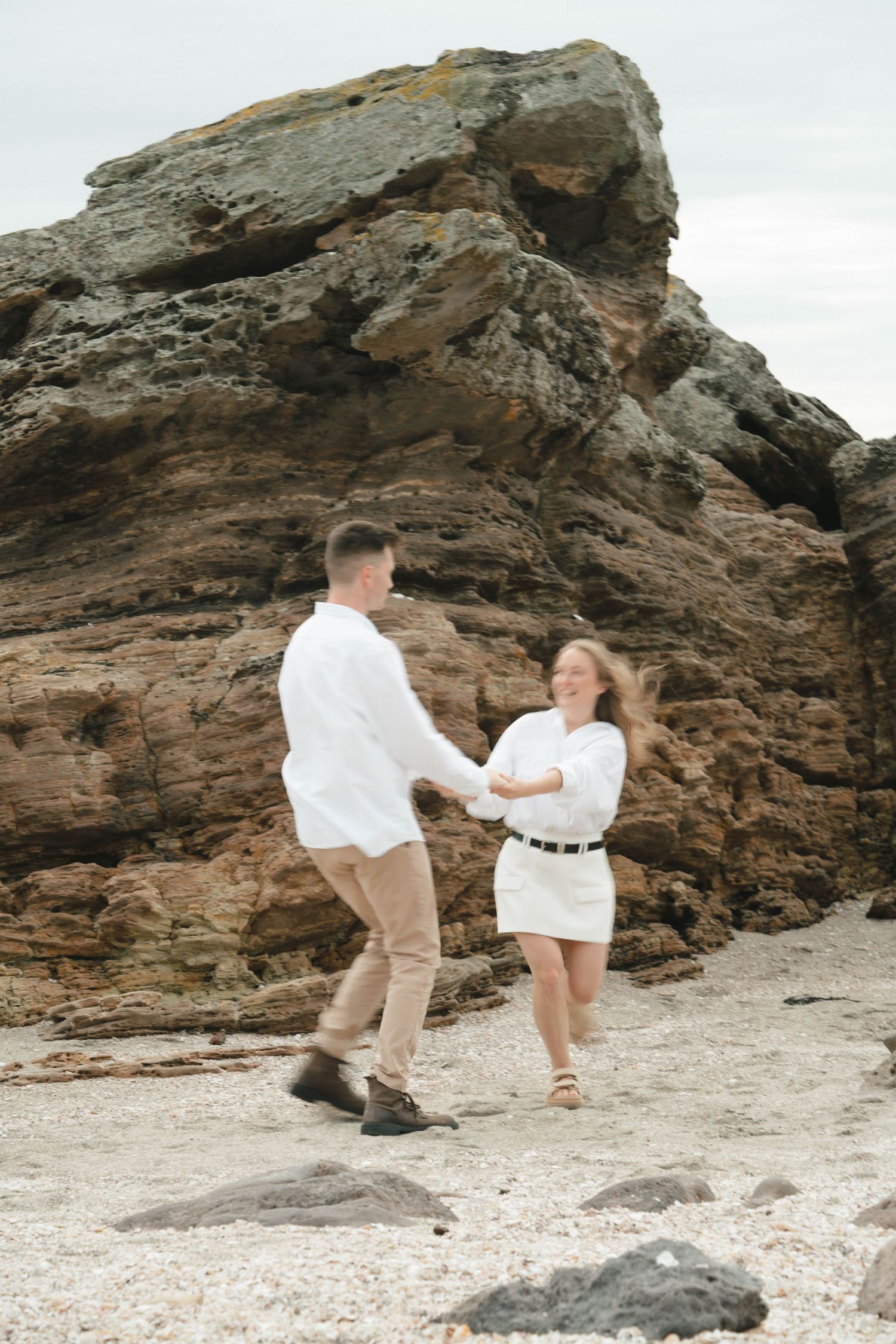 Playful couple in white clothing dancing on a beach with dramatic rocky formations in background.