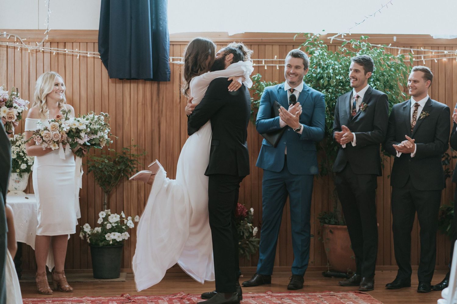 Wedding guests stand in a row against a rustic wooden wall decorated with floral arrangements.