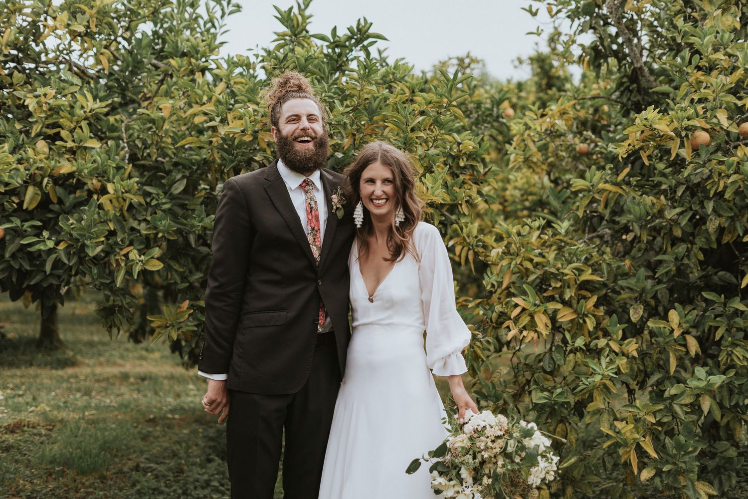 A couple poses together in a sunny orchard with green trees and foliage creating a natural outdoor backdrop.