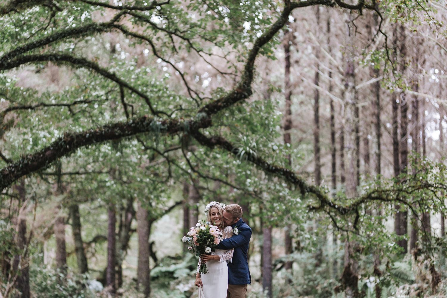 Wedding guests gather for an outdoor wedding ceremony surrounded by tall trees in a lush forest setting.