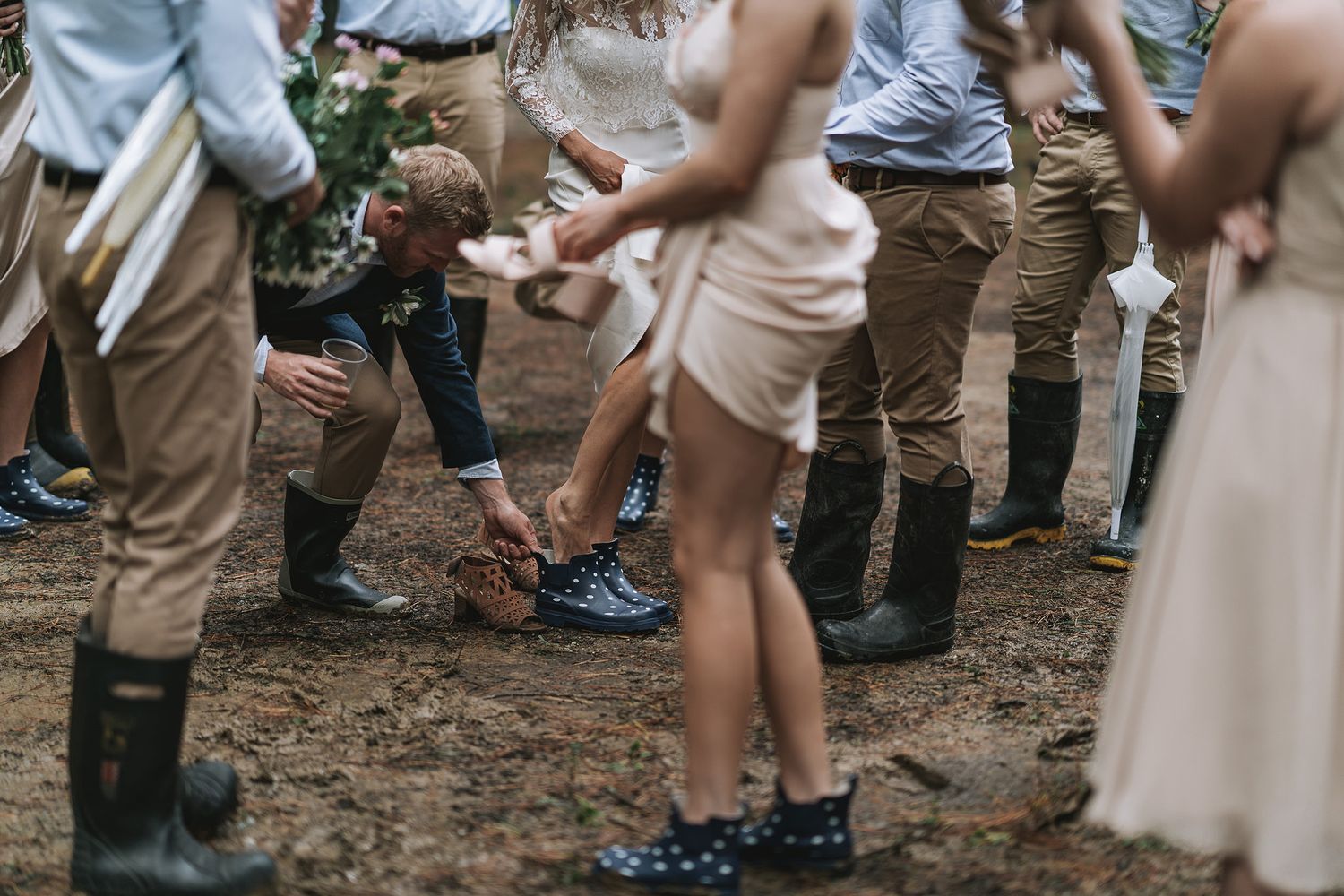 Wedding guests dance with joy at a rustic outdoor reception on dirt ground wearing formal attire.