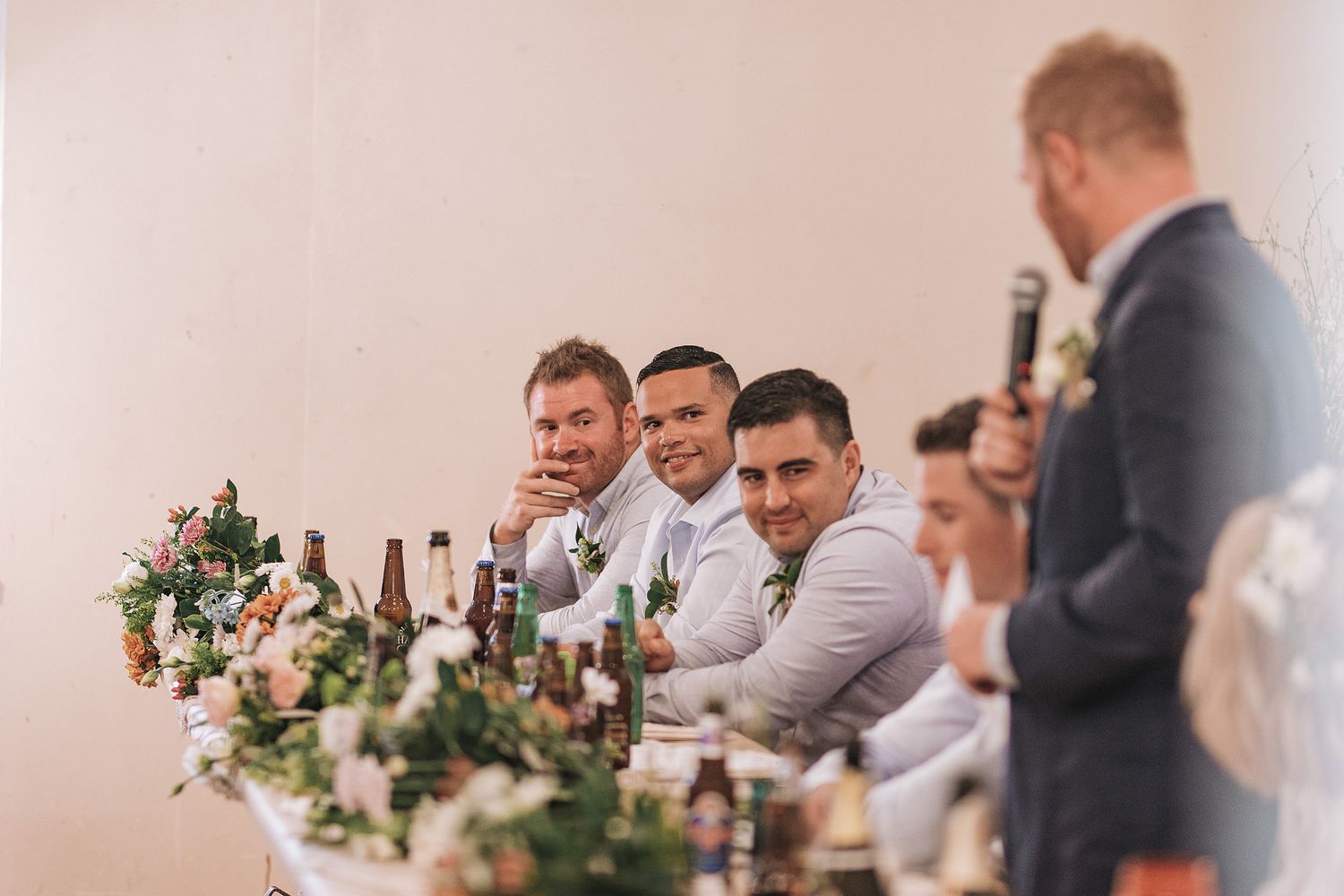 Wedding reception guests seated at the head table with floral centerpieces and wine bottles.
