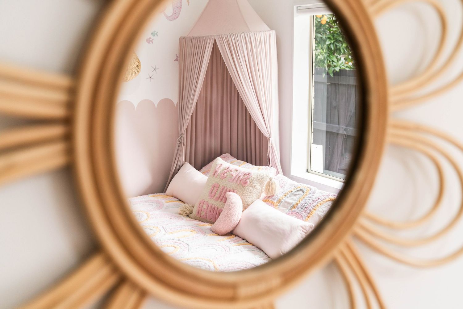 Round sunburst mirror reflecting a pink bedroom with canopy and plush bedding.