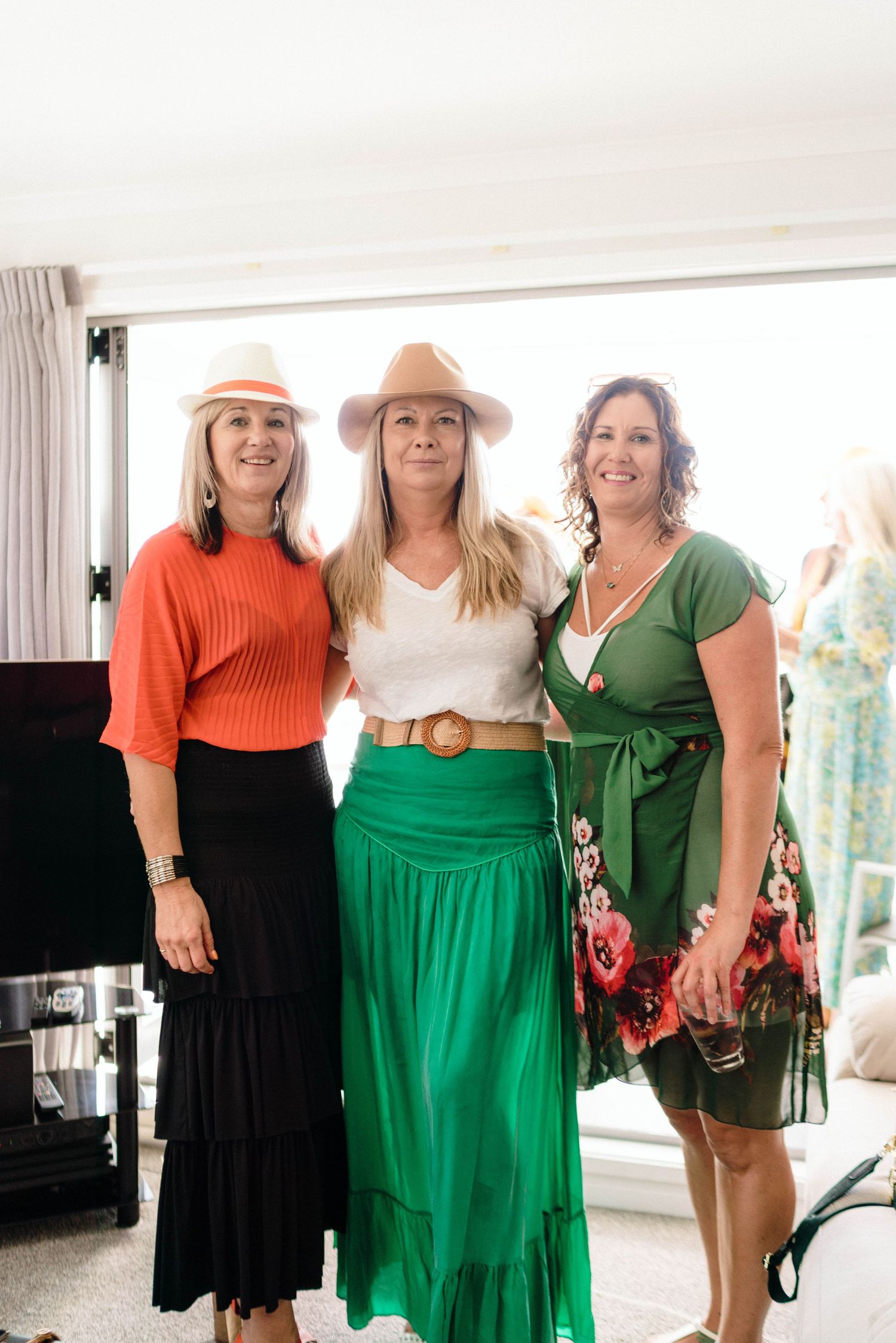 Three women in stylish outfits including orange top, white blouse with green skirt, and green dress pose together indoors.