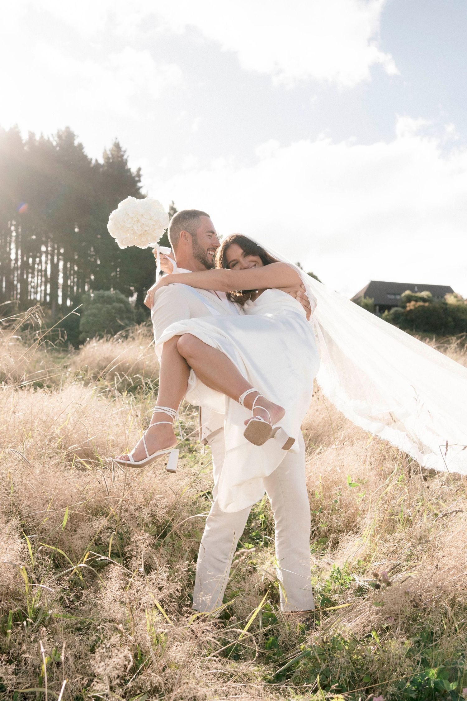A couple in all white attire dancing in a sunlit field with tall grass.