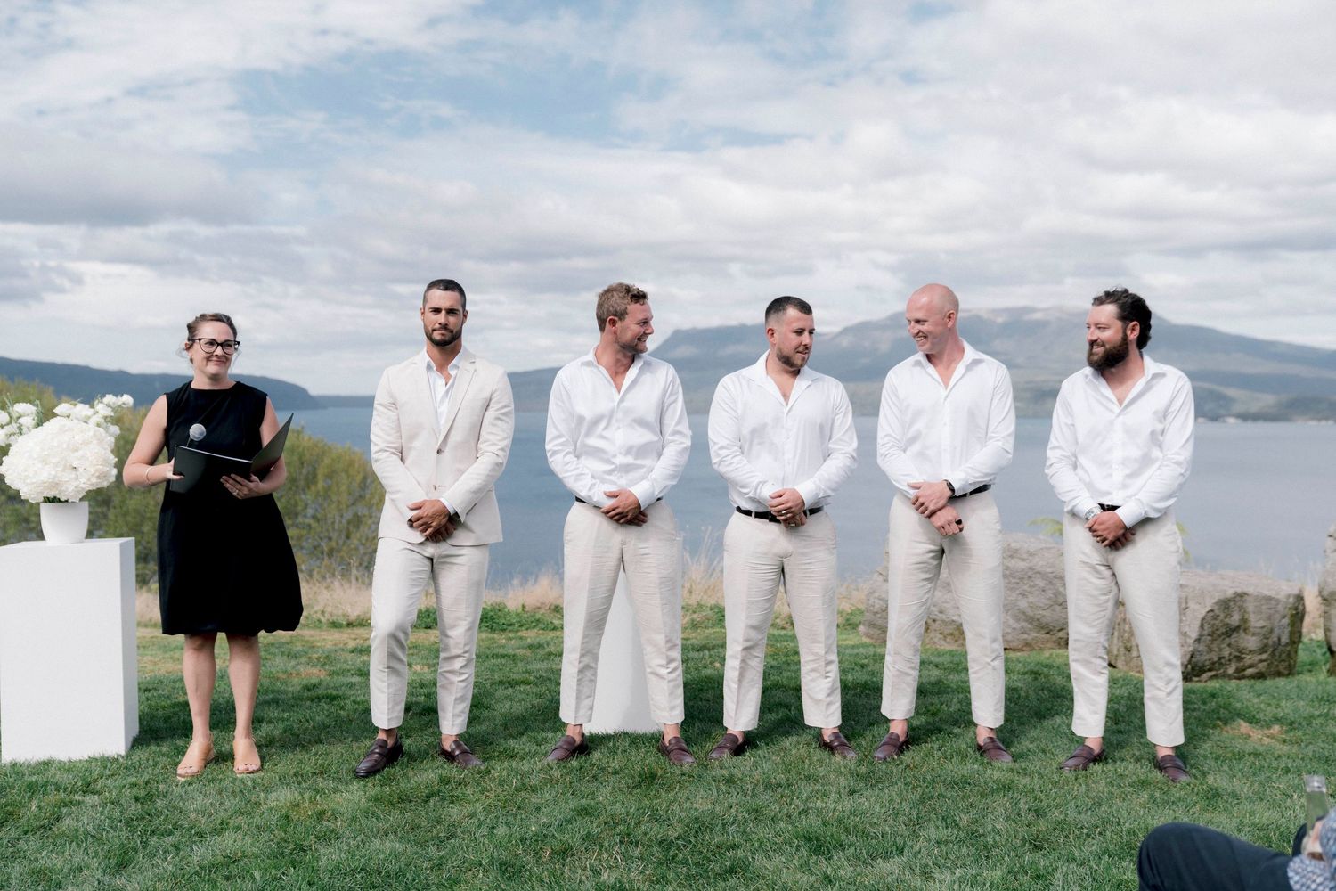 Wedding party members lined up in white attire on a cliff overlooking a scenic lake view.