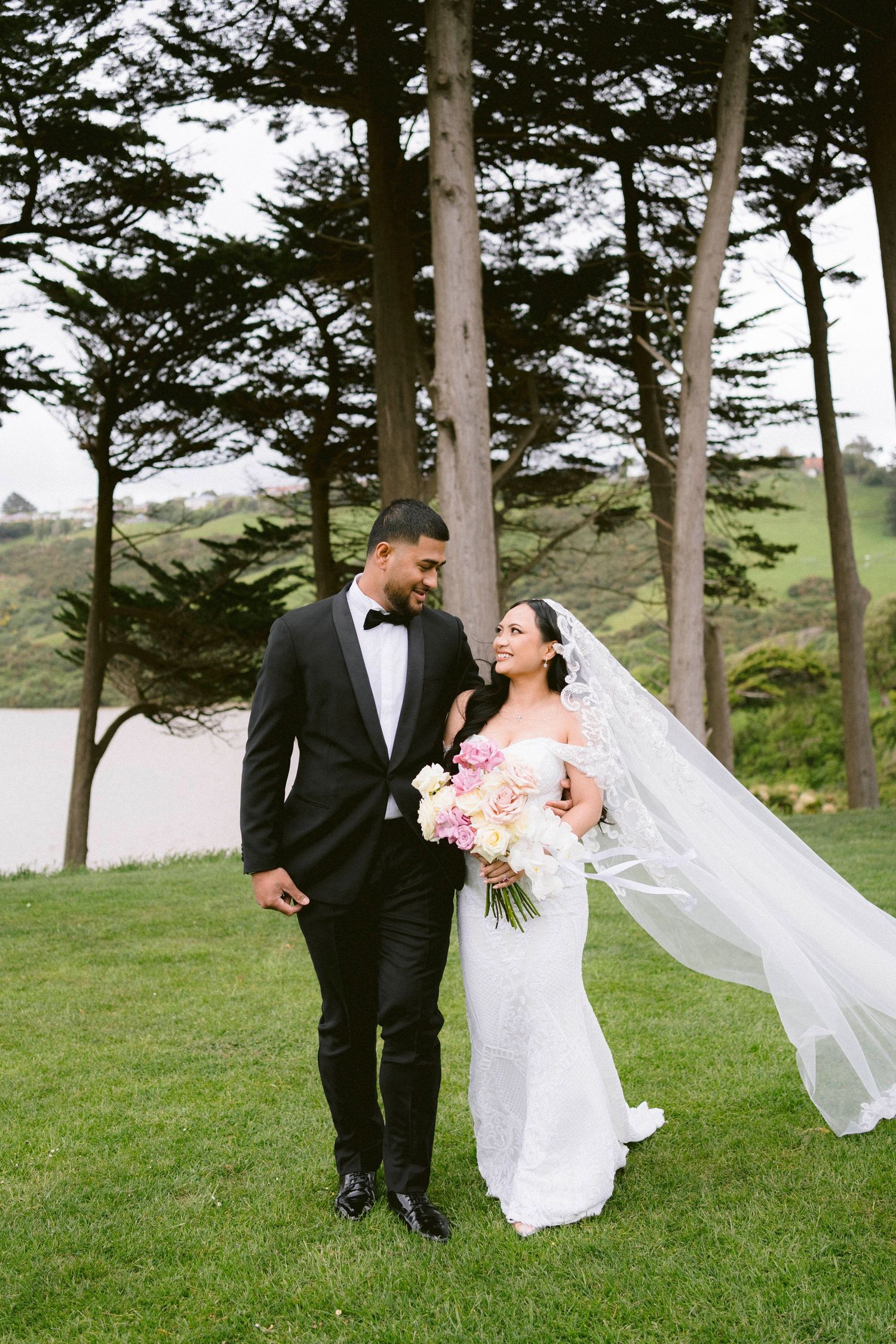 Romantic wedding couple walks together on grassy hillside with tall pine trees and flowing veil in the wind.