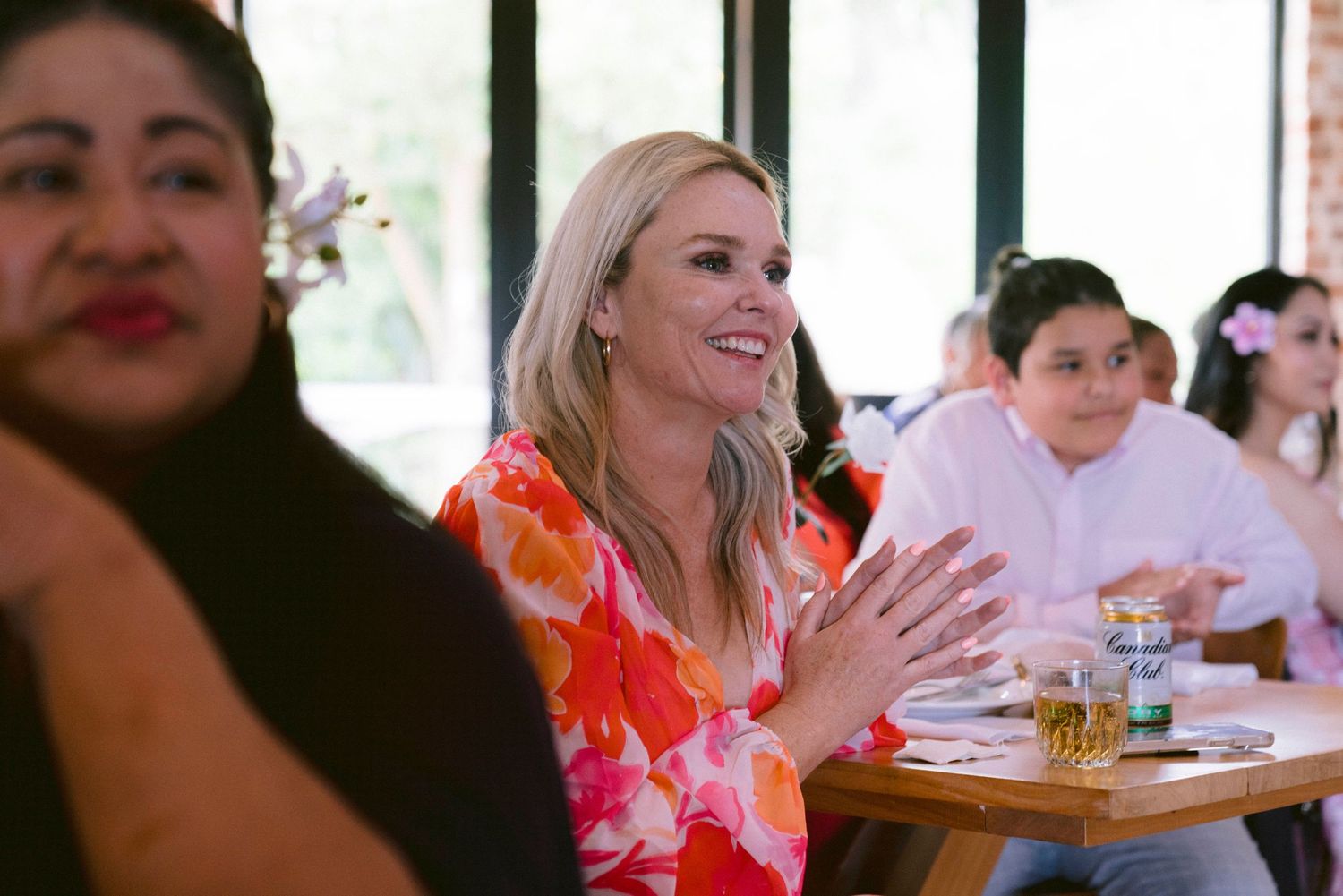 Wedding guests sit at tables during a reception celebration while clapping and smiling.