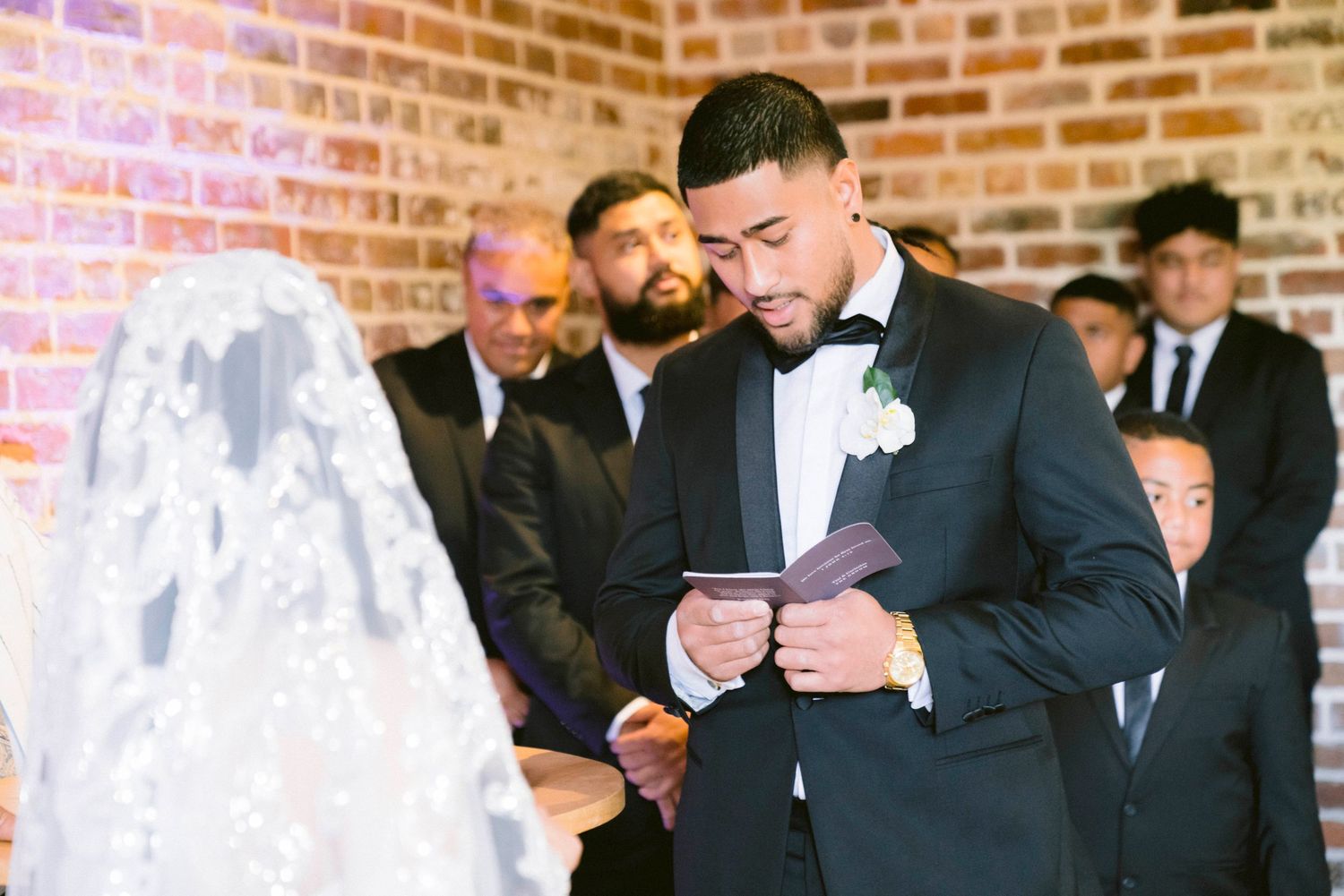 A groom reads his vows during an indoor wedding ceremony against an exposed brick wall backdrop.